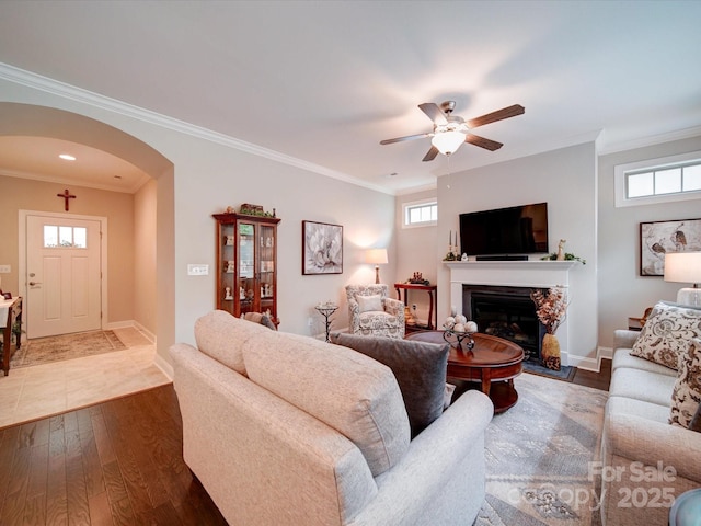 living room with a fireplace with raised hearth, a ceiling fan, arched walkways, a wealth of natural light, and dark wood-style flooring