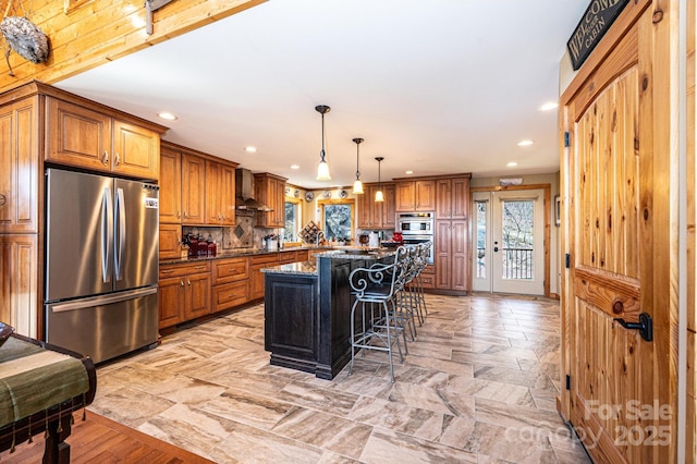 kitchen featuring recessed lighting, brown cabinetry, wall chimney range hood, and appliances with stainless steel finishes