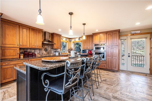 kitchen with a center island, brown cabinets, stainless steel double oven, wall chimney exhaust hood, and recessed lighting