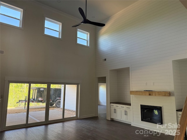 unfurnished living room with a ceiling fan, a glass covered fireplace, a high ceiling, ornamental molding, and dark wood-type flooring