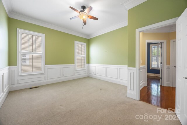 unfurnished room featuring a wainscoted wall, visible vents, a ceiling fan, crown molding, and carpet flooring