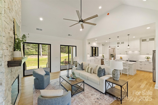 living room featuring ceiling fan, visible vents, a stone fireplace, and high vaulted ceiling