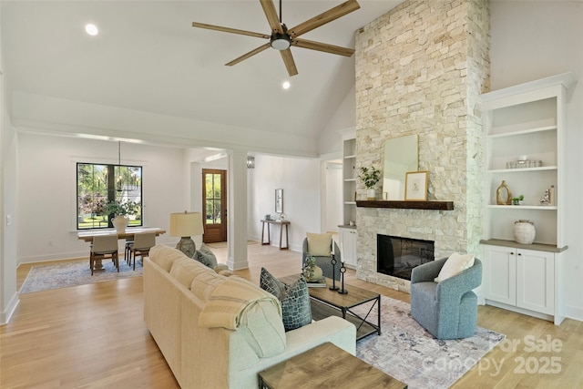 living area featuring high vaulted ceiling, a ceiling fan, a stone fireplace, light wood-style flooring, and ornate columns