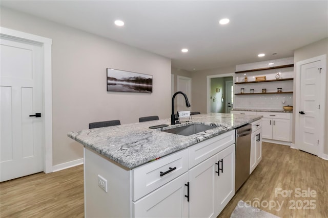 kitchen featuring a sink, stainless steel dishwasher, light wood-style flooring, open shelves, and an island with sink