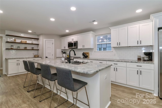 kitchen with stainless steel microwave, backsplash, a sink, white cabinets, and a kitchen island with sink