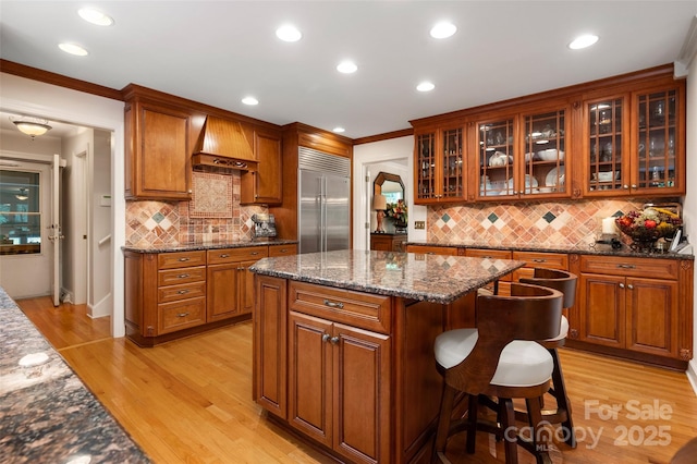 kitchen with stainless steel built in refrigerator, custom exhaust hood, light wood finished floors, backsplash, and a kitchen island