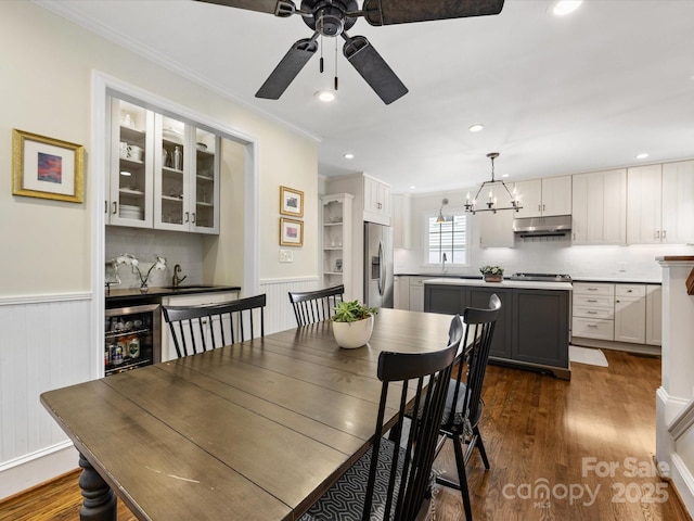 dining area with wainscoting, ornamental molding, ceiling fan with notable chandelier, dark wood-type flooring, and wine cooler