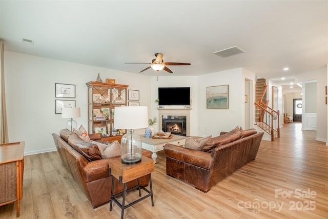 living room featuring stairway, light wood finished floors, visible vents, a glass covered fireplace, and ceiling fan