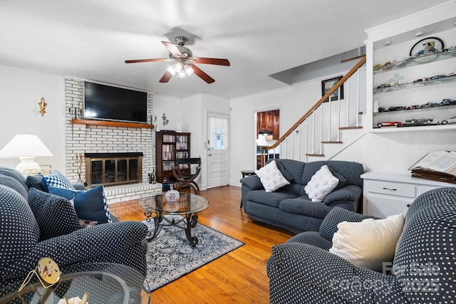 living room featuring a brick fireplace, stairs, ceiling fan, and wood finished floors