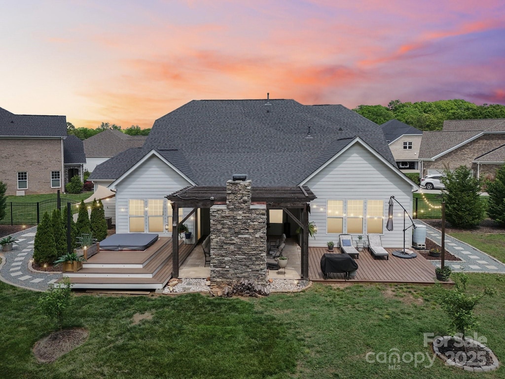 back of house at dusk with a pergola, fence, a wooden deck, a lawn, and a shingled roof