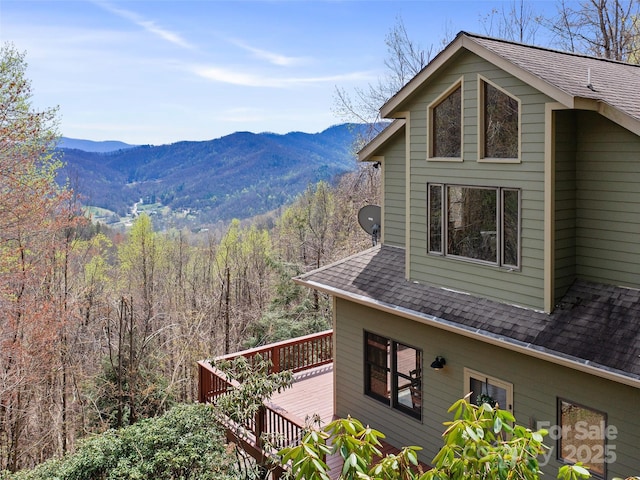 view of side of home featuring a deck with mountain view, a shingled roof, and a view of trees