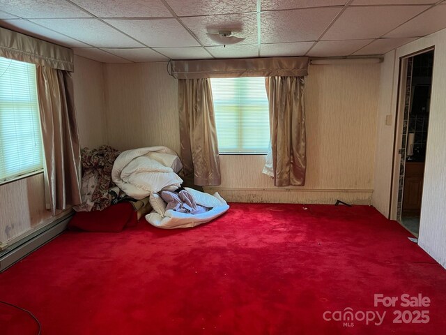 carpeted bedroom featuring wooden walls, a baseboard heating unit, and a drop ceiling