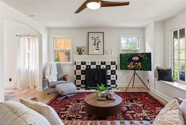 living area featuring wood finished floors, arched walkways, a ceiling fan, baseboards, and a tiled fireplace