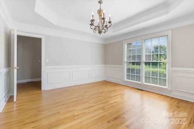 spare room with a wainscoted wall, a raised ceiling, ornamental molding, light wood-style flooring, and a notable chandelier
