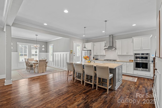 309 Catawba Ave, Davidson, NC 28036 - kitchen with wall chimney range hood, appliances with stainless steel finishes, a center island with sink, a breakfast bar, and dark wood-style flooring