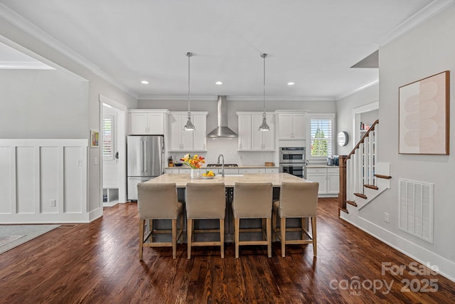 309 Catawba Ave, Davidson, NC 28036 - kitchen featuring wall chimney range hood, appliances with stainless steel finishes, a center island with sink, a breakfast bar area, and crown molding