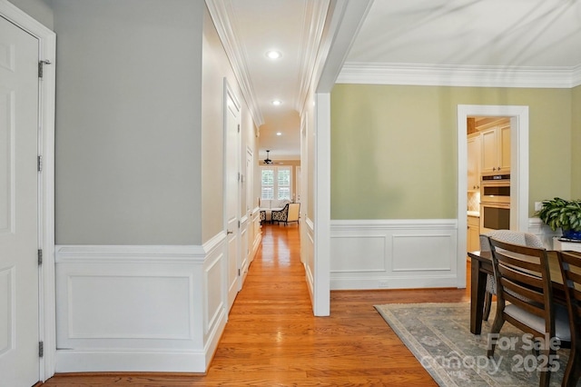 corridor with light wood-type flooring, a wainscoted wall, crown molding, and a decorative wall