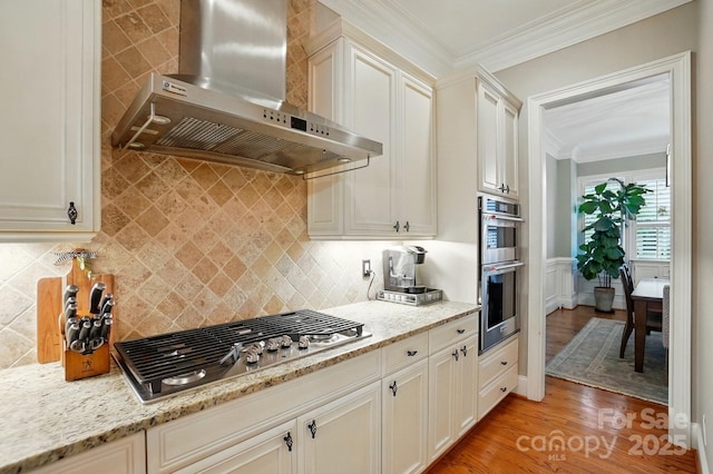 kitchen featuring appliances with stainless steel finishes, light wood-type flooring, light stone countertops, ornamental molding, and wall chimney exhaust hood