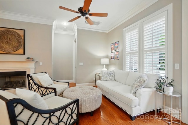 living area with wood finished floors, ornamental molding, ceiling fan, a glass covered fireplace, and baseboards