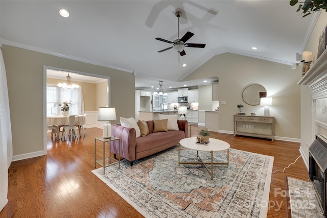 living room with vaulted ceiling, crown molding, ceiling fan with notable chandelier, a fireplace with flush hearth, and light wood-style flooring