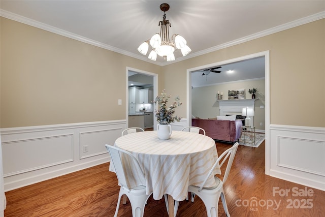dining area featuring a wainscoted wall, ceiling fan with notable chandelier, wood finished floors, crown molding, and a fireplace
