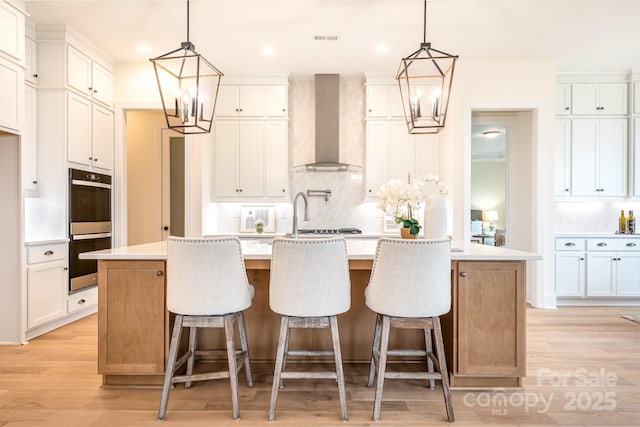 kitchen featuring backsplash, double oven, light wood finished floors, a notable chandelier, and wall chimney exhaust hood