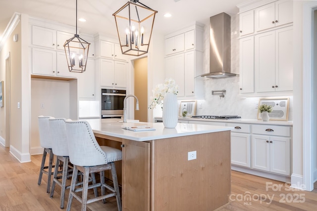 kitchen with wall chimney range hood, an inviting chandelier, light wood-style floors, light countertops, and an island with sink