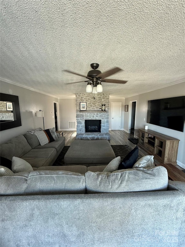 living area featuring wood finished floors, visible vents, a stone fireplace, ornamental molding, and ceiling fan