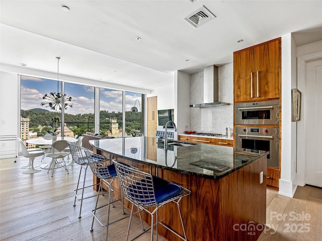7 Patton Ave Unit 1503, Asheville, NC 28801 - kitchen with wall chimney exhaust hood, appliances with stainless steel finishes, brown cabinets, light wood finished floors, and a kitchen island with sink
