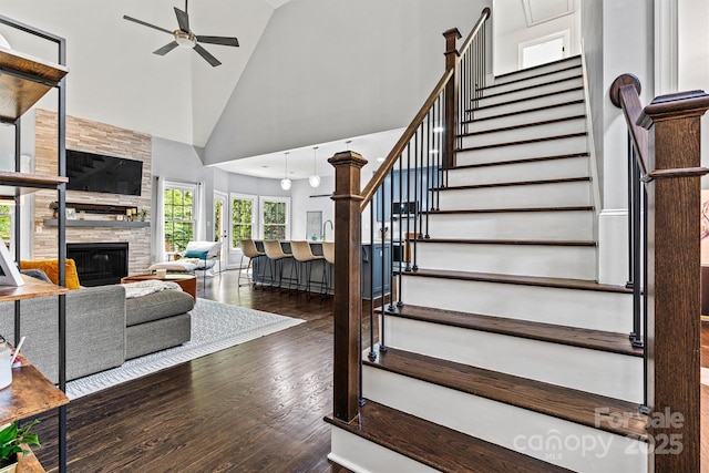 stairs with a stone fireplace, wood finished floors, high vaulted ceiling, and ceiling fan