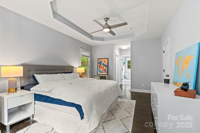 bedroom featuring a tray ceiling, dark wood-style flooring, baseboards, ceiling fan, and ensuite bathroom