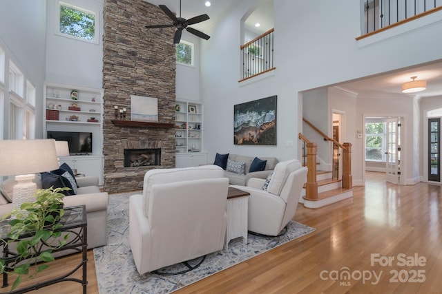 living room with a ceiling fan, a stone fireplace, stairway, light wood-style floors, and a high ceiling
