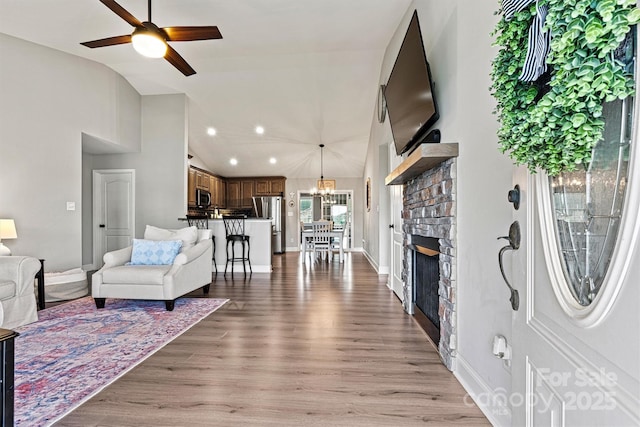 living area featuring light wood-style floors, vaulted ceiling, ceiling fan with notable chandelier, a fireplace, and baseboards