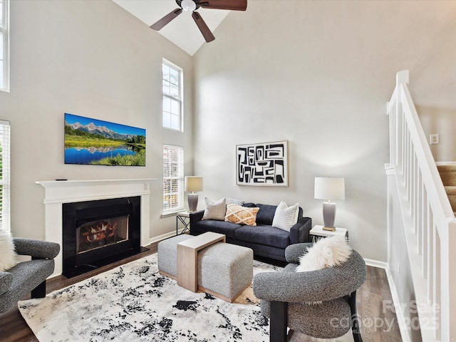 living area featuring high vaulted ceiling, wood finished floors, ceiling fan, stairway, and a glass covered fireplace