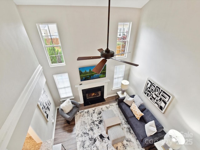 living area featuring wood finished floors, a glass covered fireplace, a towering ceiling, healthy amount of natural light, and ceiling fan