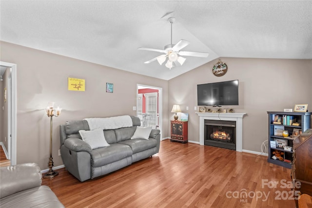 living area with lofted ceiling, wood finished floors, baseboards, a lit fireplace, and ceiling fan