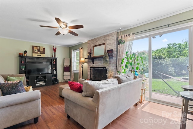 living room with a ceiling fan, crown molding, a brick fireplace, and wood finished floors