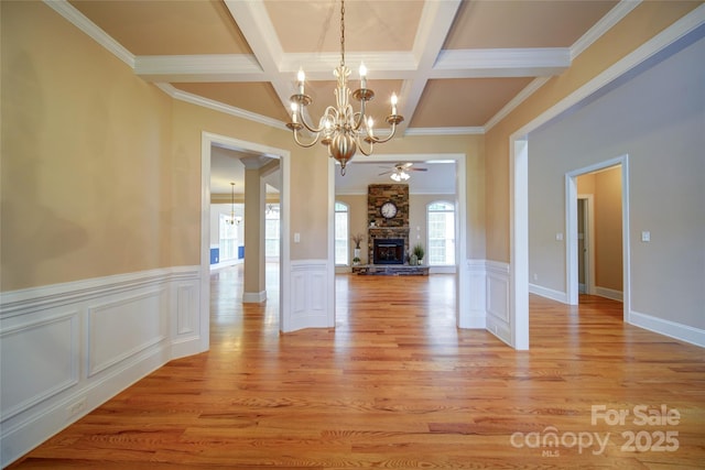 unfurnished dining area with a stone fireplace, ceiling fan with notable chandelier, beamed ceiling, light wood finished floors, and coffered ceiling