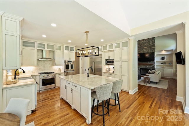kitchen with a sink, wall chimney range hood, high quality appliances, a stone fireplace, and light wood-style floors