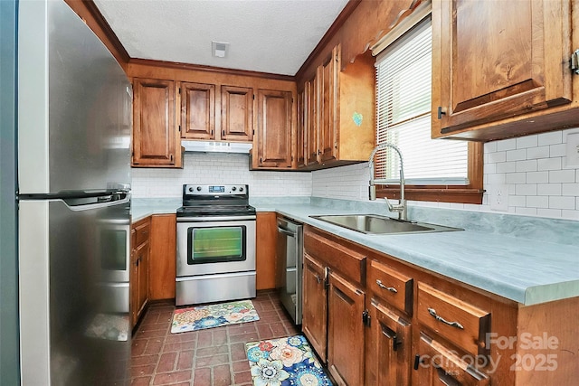 kitchen featuring a sink, brown cabinetry, stainless steel appliances, under cabinet range hood, and brick patterned floor