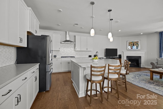 kitchen featuring open floor plan, dark wood-style floors, wall chimney range hood, a kitchen bar, and white cabinetry