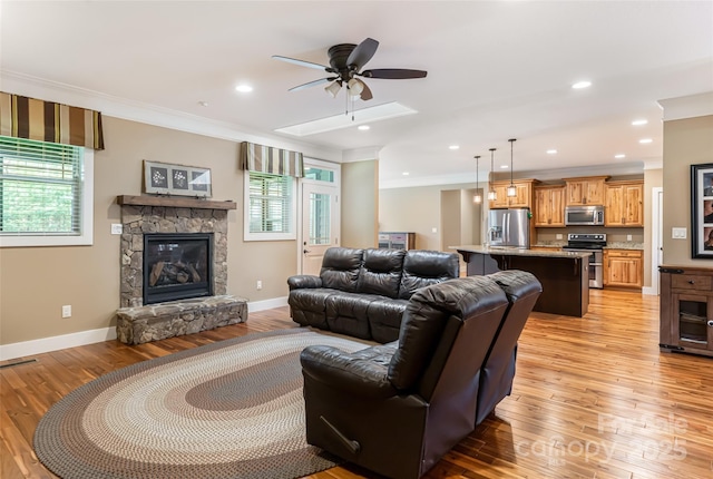 living area with ornamental molding, light wood-style floors, baseboards, a stone fireplace, and a ceiling fan