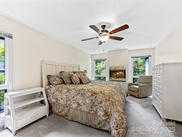 carpeted bedroom featuring a glass covered fireplace, ceiling fan, a textured ceiling, and multiple windows