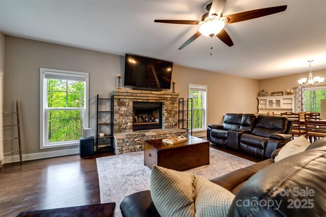 living room with baseboards, a stone fireplace, ceiling fan with notable chandelier, and dark wood-type flooring