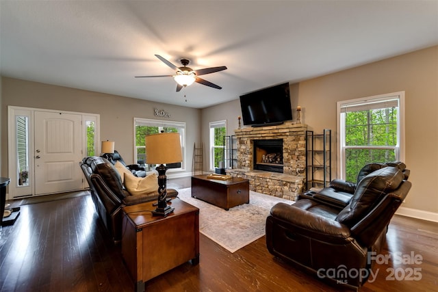 living room with dark wood-style flooring, a ceiling fan, baseboards, and a stone fireplace