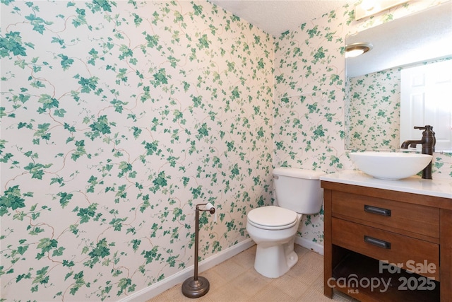 bathroom with a textured ceiling, vanity, baseboards, toilet, and wallpapered walls