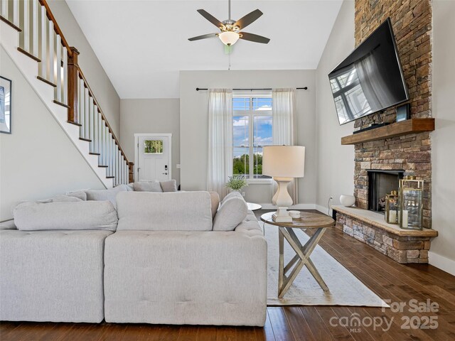 living area with stairs, a stone fireplace, ceiling fan, wood-type flooring, and baseboards
