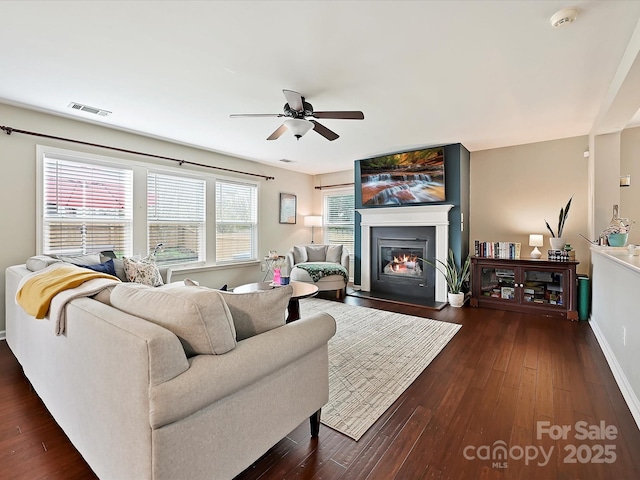 living room featuring dark wood-type flooring, a glass covered fireplace, visible vents, and ceiling fan