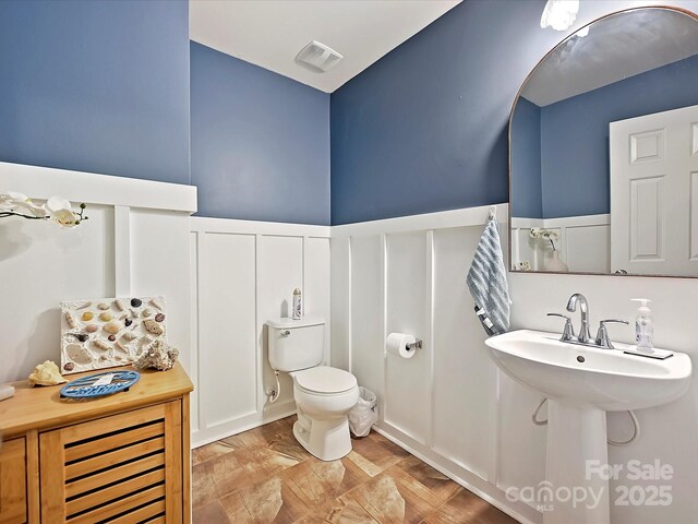 half bathroom with a decorative wall, visible vents, toilet, and wainscoting