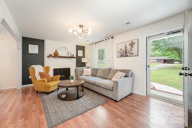 living room featuring plenty of natural light, light wood finished floors, a brick fireplace, and a notable chandelier